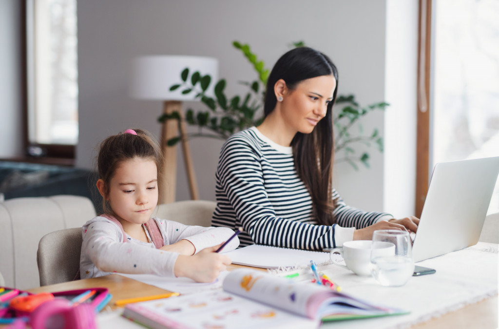 Little girl doing her homework beside her mother who is working